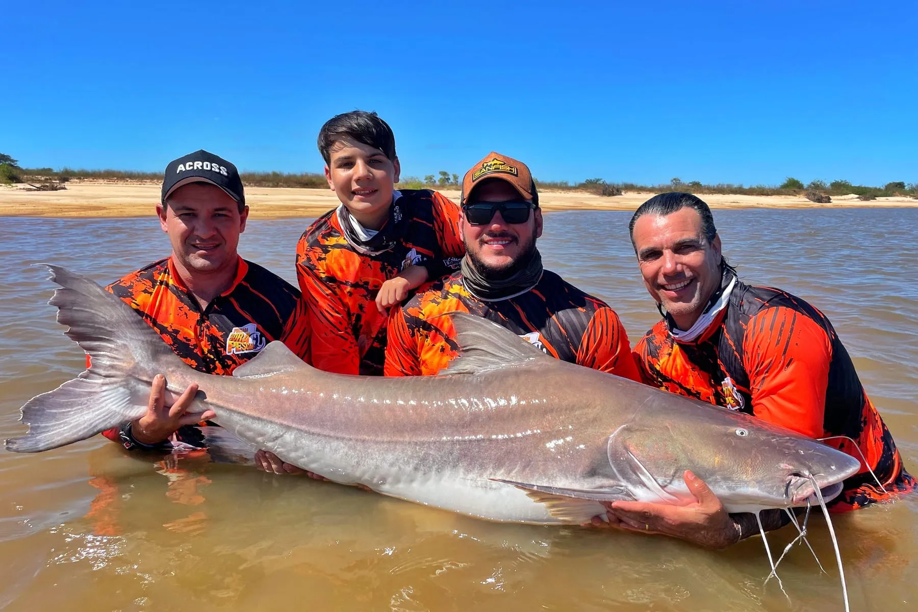Quatro pescadores, entre eles um menino, seguram um peixe de couro enorme dentro do rio
